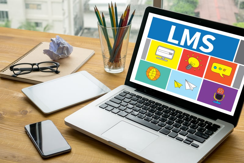 Laptop displaying a learning management system dashboard beside a tablet, smartphone, notebook, and pencils.